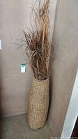 Front angle view of a tall woven cylindrical basket filled with dried grass decorations placed against a beige wall.