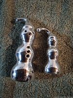 Front view of two shiny aluminum snowman sculptures with curved top hats, showing their smooth reflective surfaces and button details.