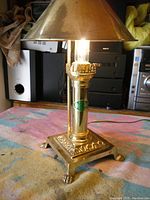 Full view of brass table lamp with conical shade on and lit bulb showing polished brass column and base with claw feet.