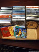 Box of mixed music CDs including rock artists and Christmas albums in jewel cases.