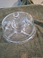 Clear glass dome cake cover with knob handle, sitting on green textured fabric background.