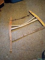 Vintage cross cut buck saw laid flat on carpeted floor, showing wooden frame and metal saw blade with teeth and tensioning wire.