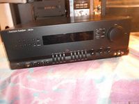 Black vintage Harman Kardon AM 25a receiver amplifier showing front panel with buttons, sliders, and display screen in room setting.