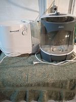 Two humidifiers side by side on a green towel: white Honeywell with control dial on left, grey Sunbeam on right with transparent water tank and power cords.