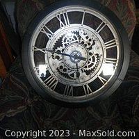 Front view of large gear clock showing black frame, Roman numerals, and internal metal gears.