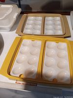 Two vintage Tupperware deviled egg carriers, one beige and one yellow, both with two plastic trays featuring 12 egg slots each.