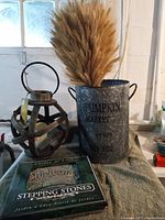 Overall view of cylindrical galvanized metal pail labeled 'PUMPKIN MARKET No. 102' containing a wheat bundle, adjacent to a lantern and a garden stepping stone.