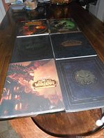 Six World of Warcraft books arranged on wooden table, five hardcover and one softcover, in mint condition with packaging on five.