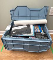 Closed-top blue plastic tote on wood floor showing variety of art supplies inside