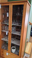 Front-right angled view of wooden cabinet showing glass doors, glass shelves with glassware inside, and two drawers at base.