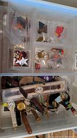 Photo showing assortment of watches with leather and metal bands placed in a plastic container, alongside various costume jewelry pieces in plastic compartments.