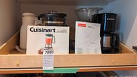 Overall view of lot items on a wooden shelf showing Cuisinart blender in box, Cuisinart drip coffee maker, Bodum coffee grinder, and two white thermoses.