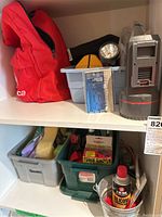 Shelf with red emergency kit bag, plastic bins containing flashlight, electrical cords, and packaged hardware items, and silver portable heater with purple base.