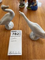 Pair of white Lladro porcelain geese figurines on wooden surface with MaxSold lot card.