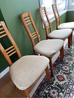 Four wooden dining chairs with ladder-style backs and beige fabric seats arranged in a line along a green wall and on a patterned rug.
