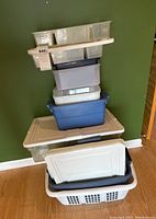 Stack of various plastic storage boxes and totes seen from front. Includes clear compartment tray, blue, gray, and white containers with lids and laundry basket at bottom.