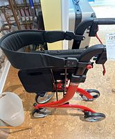 Side view showing red metal frame, black padded seat and four wheels.