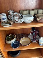 Shelves with pottery items including pitchers with brown decorative designs, votive candle holders, a white vase with side handles, a pottery candlestick holder, and various bowls and cups.