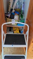 White metal step stool with black treads on two steps in a closet with other cleaning supplies visible behind