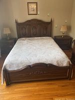 Front view of queen size wooden bed frame with headboard, bedspread, and two matching nightstands with lamps.
