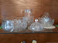 Full view of four crystal items on glass shelf: large bowl, pitcher, covered plate, and handled bowl