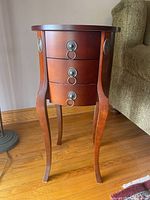 Front view of the reddish-brown wooden circular end table showing three small drawers with brass ring-pull handles and curved legs.