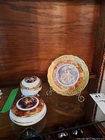 Overall view of the decorative plate on a metal stand next to two Limoges porcelain boxes on a wooden shelf