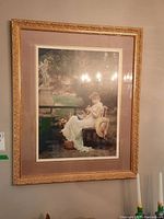 Framed print showing a woman in white dress reading a book outdoors in garden setting, enclosed in gold ornate frame.