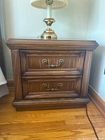 Pair of wooden side tables with two drawers, showing full front angle of one table with a lamp on top.
