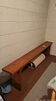 Side view of long, narrow solid wood bench against a wall on a brown floor.
