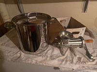 Photo showing both the large stainless steel stock pot with lid and the heavy counter-mounted metal meat grinder with wooden handle placed side by side on a cloth-covered surface.
