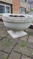 Close-up photo of one cement urn planter showing its round bowl shape with a squared pedestal base. The planter is filled with soil and placed on a brick patio.