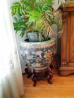 Full view showing vintage large ceramic planter with colorful bird and floral design sitting on ornate wooden stand, live plant in plastic pot inside planter
