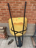 Front view of yellow wheelbarrow showing metal tray with rust and black handles against brick wall