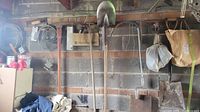 Photo showing pick axe, shovels, drain snake, hand trowel hanging on garage wall with brick background