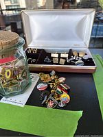 Assorted cufflinks and pins visible in and around a white satin-lined box with multiple metal designs and patterns.