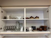 Wide view of kitchen cabinet shelves containing tea cups, saucers, spice rack, glass jars, white ceramic gravy boats, and small brown ceramic pots.