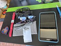 Lot contents on a black surface including four pairs of glasses, red watch, metal key and Acer tablet.