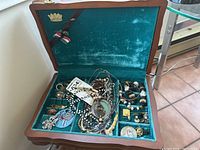 Open wooden jewellery box with green velvet lining showing various costume jewellery such as bracelets, pins, and necklaces inside compartments.