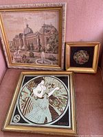 Three framed pictures arranged on a pink surface: a large needlepoint garden scene, a smaller framed round floral design, and a framed Art Nouveau style decorative tile.