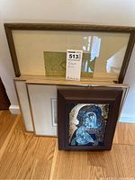 Four framed pictures leaning against wall showing a variety of artwork including a religious image in a dark frame and three frames in light colors with prints.