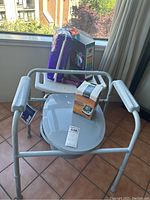 Shower seat with armrests and commode bucket seen with items on seat and tiled floor background with window and curtain.