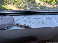 Sterling silver necklaces, 10K gold pins, and a hand mirror displayed on a windowsill with outdoor background visible.
