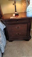 Front view of one dark brown Broyhill wood nightstand with lamp on top showing three drawers and hardware