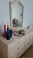 Frontal view of wicker dresser with six drawers and matching mirror above it, showing the woven wicker texture and arrangement of decorative vases and bowl on top of dresser.