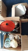 Top view of three cardboard boxes filled with mixed kitchen items including cookware with red lid, white pitchers, and metal and plastic kitchenware.