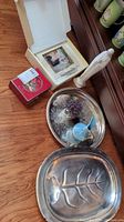 Photo showing all items including the Lenox frame in box, Waterford ornament in box, two silver-tone serving trays, Hummel figurine, and blue bird-shaped vase on wooden floor.