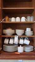 Full cabinet shelf view showing stacked plates, coffee mugs hanging, and frosted glasses on top shelf.