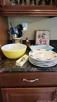 Photo showing yellow Pyrex mixing bowl, stack of six white Christmas plates with holly designs, blue Pyrex rectangular pan, ceramic utensil holder with utensils, two pieces of wall art, and decorative glass wine stopper in box.
