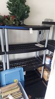 Three black plastic shelving units with silver legs, showing overall condition and size relative to nearby objects such as plastic storage bins and artificial greenery.